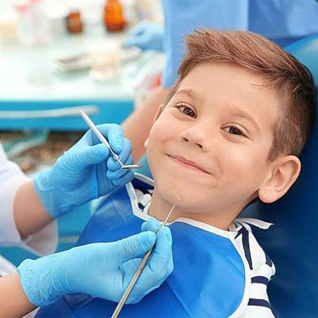 Pediatric dentist in San Diego kneeling to talk to a smiling young boy with special needs in a dental chair.